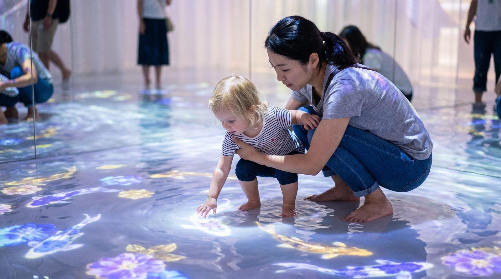 Japanese nanny helping blonde toddler explore digital koi projections in shallow water at TeamLab Planets