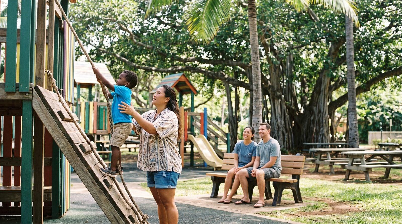 Hawaiian nanny helping toddler climb at Keiki Zoo playground while parents rest nearby